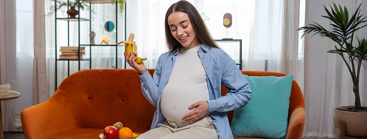 happy pregnant woman with great appetite peels eats banana fruit home healthy food concept